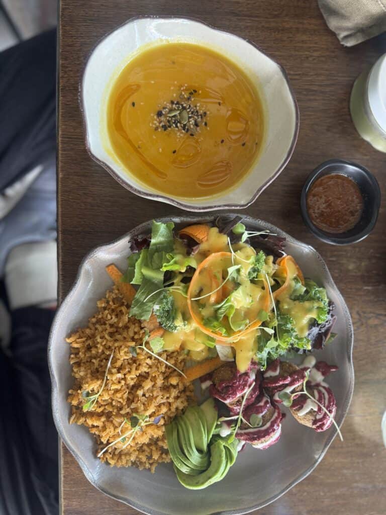Plato de ensalada variada con arroz, verduras y aderezo, acompañado de una sopa de calabaza en un bol blanco, en un ambiente de comida saludable y natural.